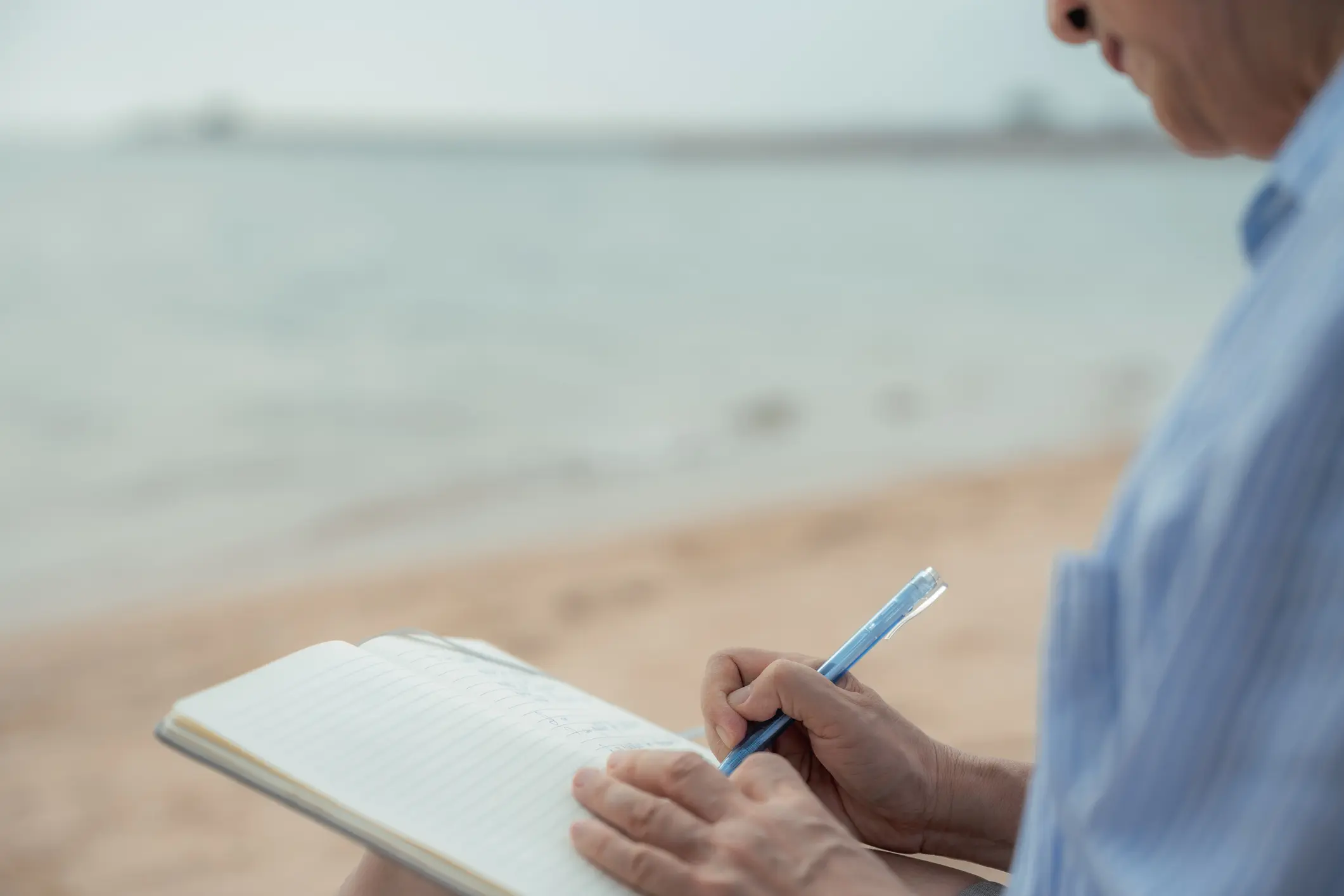 Person writing in notebook by the beach.